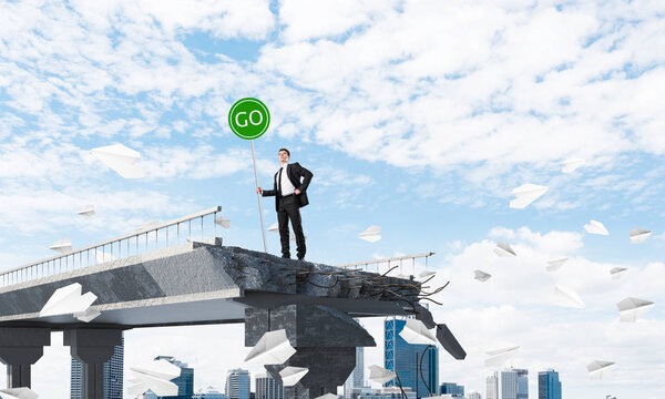 Confident businessman in suit holding green go sign while standing among flying paper planes on broken bridge with cityscape on background. 3D rendering.