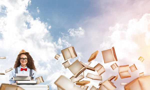 年轻和美丽的女作家在帽子和眼镜使用打字机 而坐在桌子之间的云影像的背景下的飞行书籍 — 图库照片