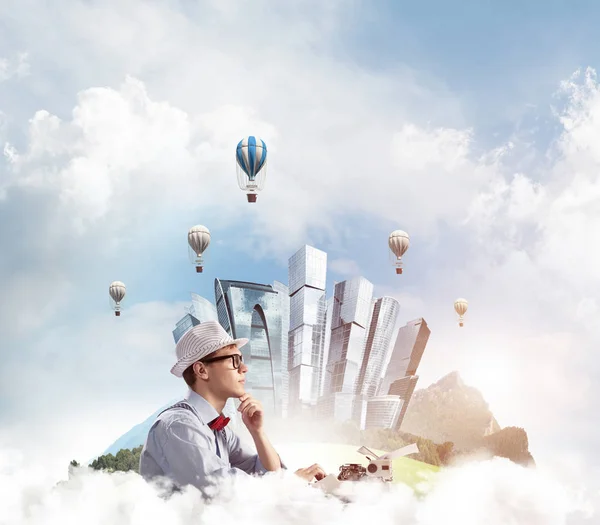 Young man writer in hat and eyeglasses using typing machine while sitting at the table with floating city and cloudy skyscape with flying aerostats on background.