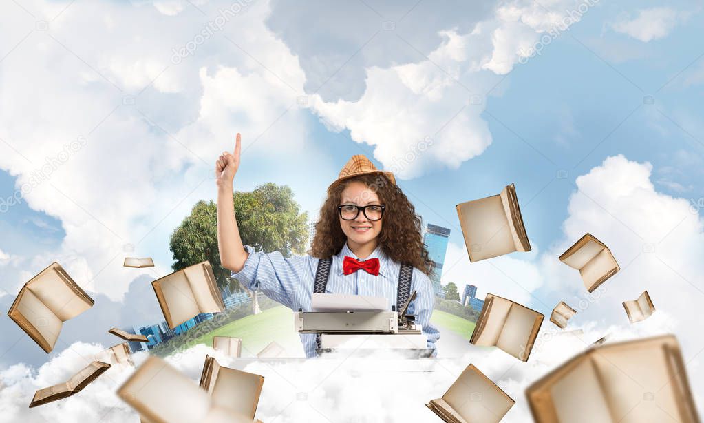 Beautiful woman writer in hat and eyeglasses using typing machine and pointing upside while sitting at the table among flying books with floating city island and cloudy skyscape on background.