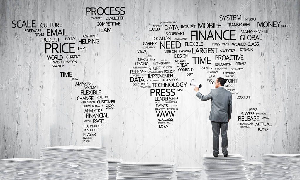 Businessman in suit standing on pile of documents with speaker in hand with business-related terms in form of world map on background. Mixed media.