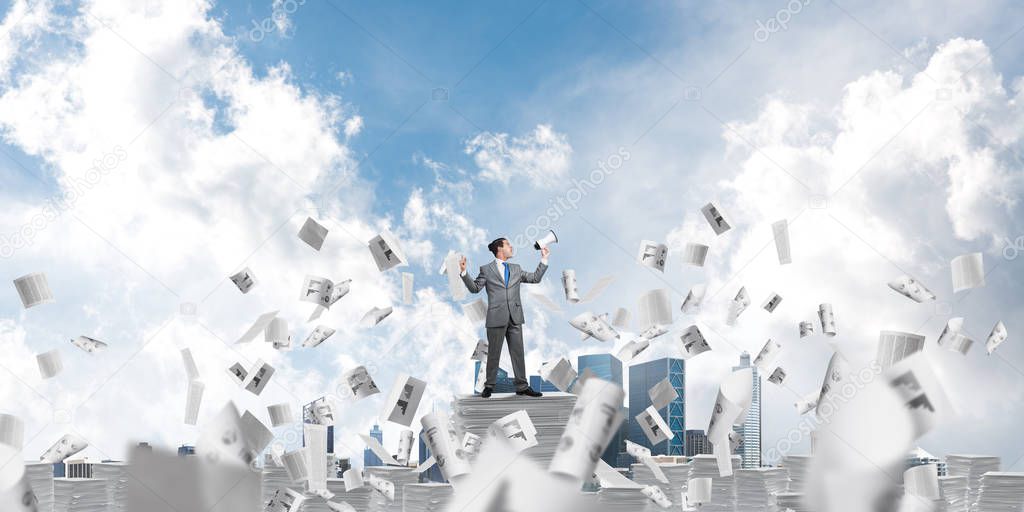 Businessman in suit standing among flying papers with speaker in hand and with skyscape on background. Mixed media.