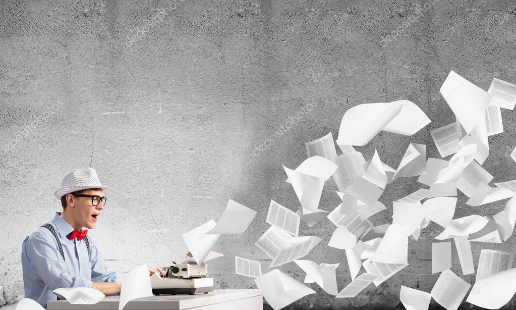 Young man writer in hat and eyeglasses using typing machine while sitting at the table among flying papers and against gray concrete wall on background.