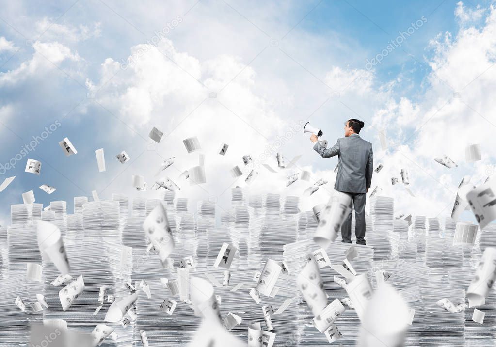 Businessman in suit standing on pile of documents among flying papers with speaker in hand with cloudly skyscape on background. Mixed media.