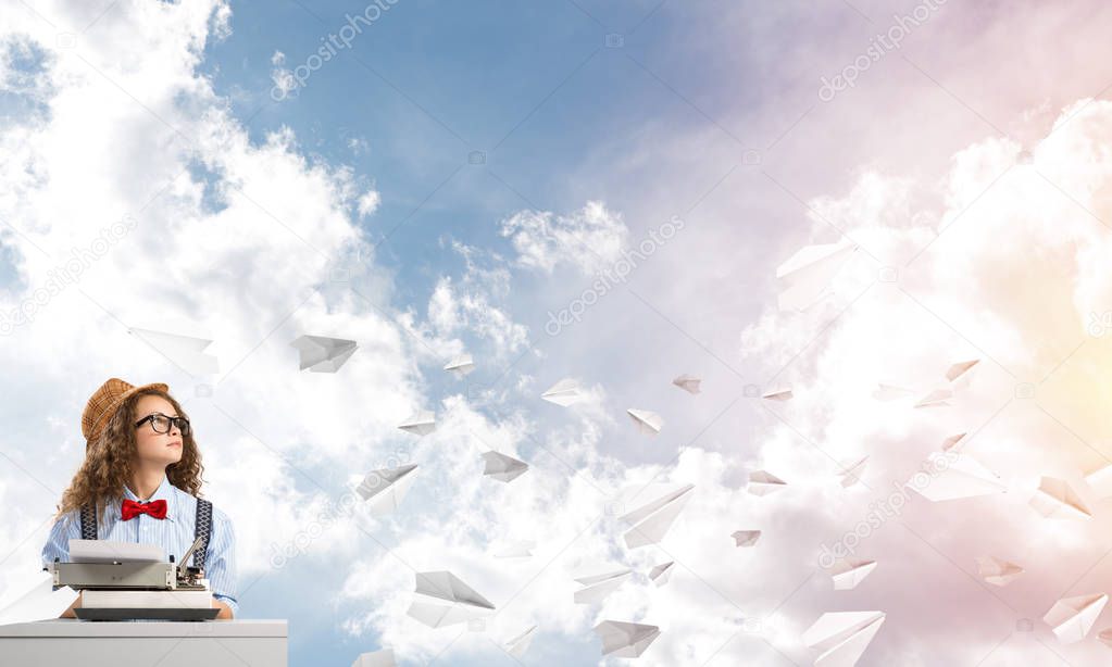 Young and beautiful woman writer in hat and eyeglasses using typing machine while sitting at the table among flying paper planes with cloudy skyscape on background.