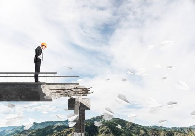 Genç takım elbiseli mühendis ve arka plan üzerinde hava ve Doğa Manzaralı kırık köprüde kağıt uçak uçan arasında duran bakıyordu kask. 3D render.