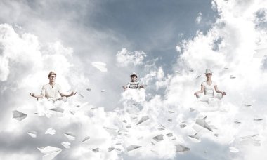 Genç aile gözlerini kapalı tutuyor ve odaklanmış görünüyor. Arka planda bulutlu bir gökyüzü ile havada uçan kağıt uçaklar arasında meditasyon yaparken..