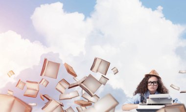 Genç ve güzel bir kadın yazar şapka ve gözlük kitaplar ile bulutlu gökyüzü arka plan üzerinde uçan arasında masadaki otururken yazarak makinasıyla.