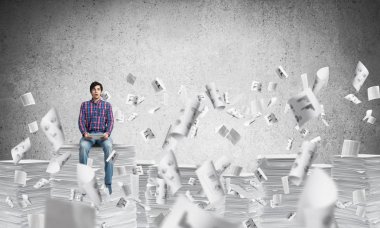 Kağıt belgeleri ile gri duvar arka plan üzerinde uçan arasında casual giyim oturan genç adam. Karışık teknik.