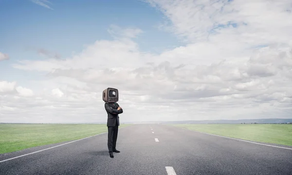 Businessman in suit with old TV instead of head keeping arms crossed while standing on the road with beautiful landscape on background.