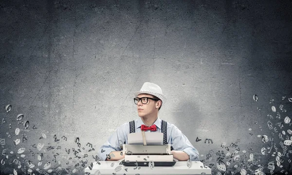 Young man writer in hat and eyeglasses using typing machine while sitting at the table among flying letters and against gray concrete wall on background.