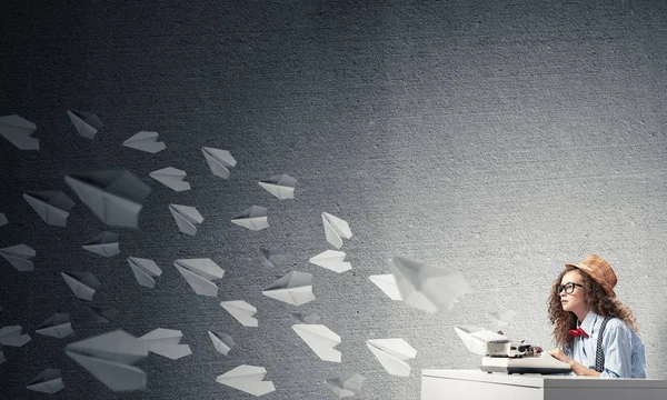 Young and beautiful woman writer in hat and eyeglasses using typing machine while sitting at the table among flying paper planes and against gray concrete wall on background.