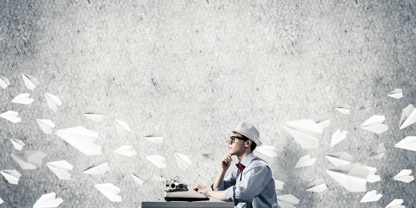 若い男の帽子と眼鏡の背景に紙飛行機を飛んでいる間 灰色のコンクリートの壁にテーブルに座ってタイピング マシンを使用しての作家 — ストック写真