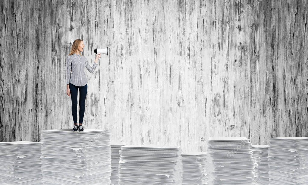 Woman in casual clothing standing on pile of documents with speaker in hand with grey wall on background. Mixed media.
