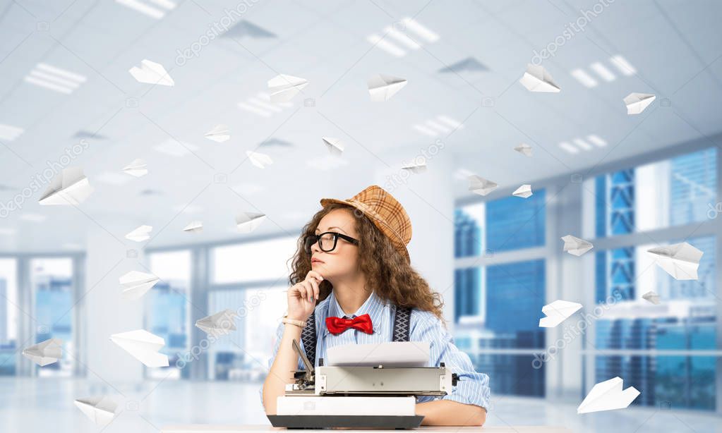 Young woman writer in hat and eyeglasses using typing machine while sitting at the table indoors among flying paper planes and with office view on background.
