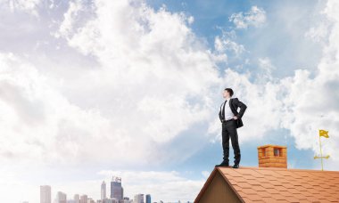 Kararlı genç işadamı evin çatısında dikiliyor ve başka tarafa bakıyor. Karışık ortam