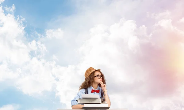 年轻和美丽的女作家在帽子和眼镜使用打字机 而坐在桌子上多云影像在后台 — 图库照片