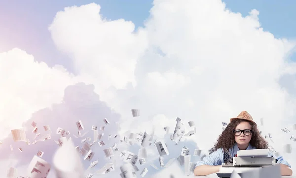 年轻而美丽的女作家在帽子和眼镜使用打字机 而坐在桌子上的飞行文件与多云的天空背景 — 图库照片