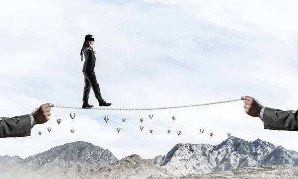 隠された脅威とサポートのシンボルとして高い山の上のロープに目隠し歩くビジネスマン 空飛ぶ風船と自然観の背景 — ストック写真