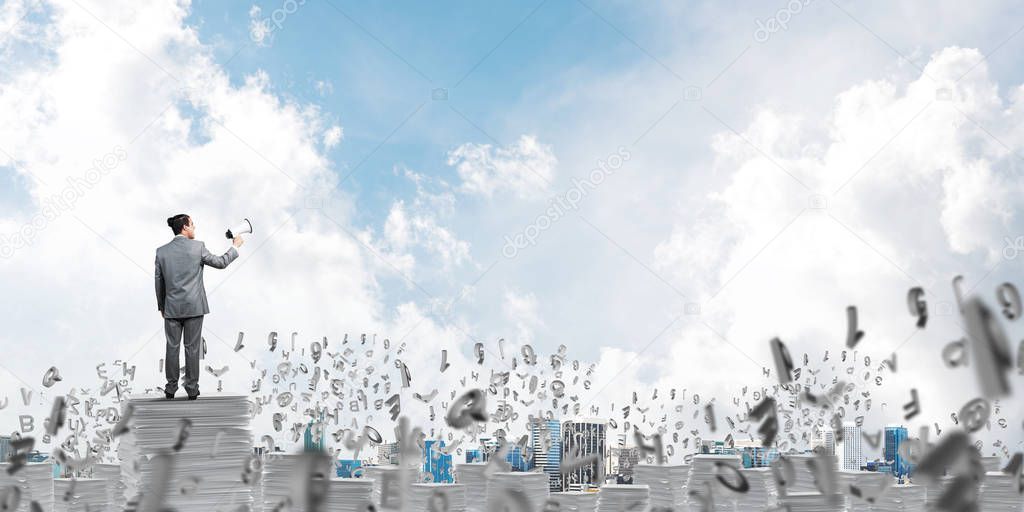 Businessman in suit standing among flying letters with speaker in hand and with skyscape on background. Mixed media.