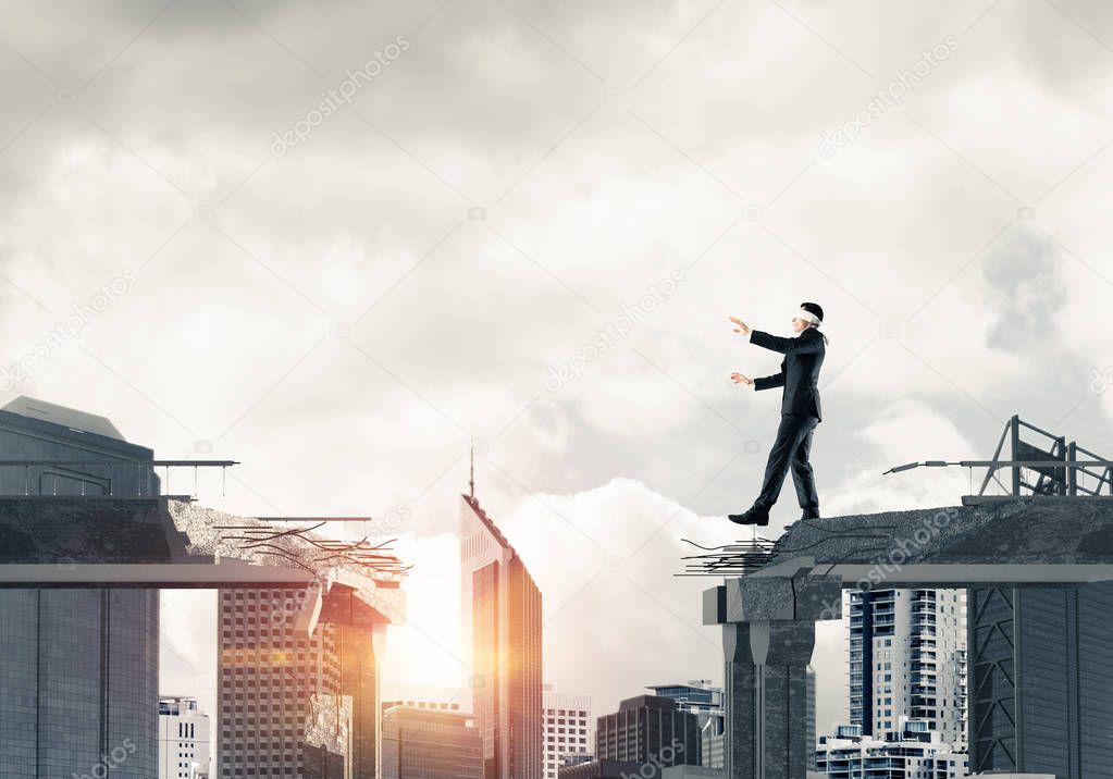 Businessman walking blindfolded on concrete bridge with huge gap as symbol of hidden threats and risks. Cityscape view with sunlight on background. 3D rendering.