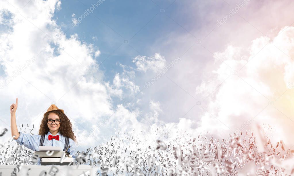 Young and beautiful woman writer in hat and eyeglasses using typing machine and pointing upside while sitting at the table among flying letters with cloudy skyscape on background.