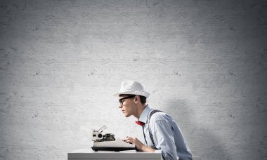 Genç yazar şapka ve gözlük arka planda gri beton duvara karşı masada otururken yazarak makinasıyla adam.