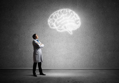 Beyaz Tıbbi üniforma silah tutmak ve arka planda gri duvara ayakta iken parlayan beyin sembolü bakarak Genç ve emin doktor.