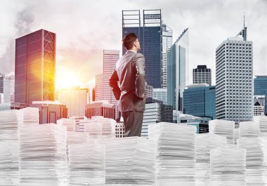 Takım elbiseli kendine güvenen genç işadamı şehir manzarası ve arka planda güneş ışığı olan bir yığın belgenin üzerinde duruyor. Karışık ortam.