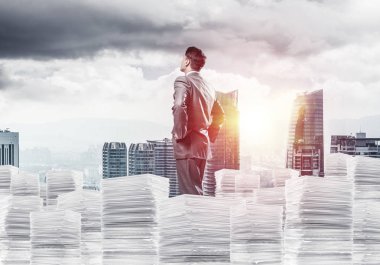 Takım elbiseli kendine güvenen genç işadamı şehir manzarası ve arka planda güneş ışığı olan bir yığın belgenin üzerinde duruyor. Karışık ortam.