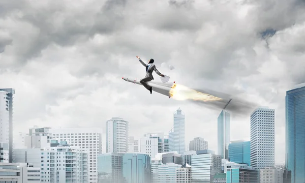 Conceptual image of young businessman in suit flying on rocket with modern cityscape with skyscrapers and blue sky on background.