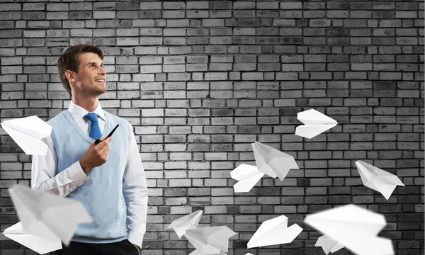 Horizontal shot of confident and young businessman in smart-casual wear smoking pipe while standing among flying paper planes and against gray dark wall on background.