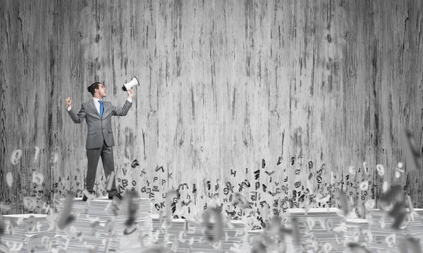 Businessman in suit standing among flying letters with speaker in hand with grey wall on background. Mixed media.