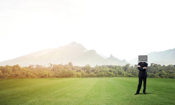 Businessman in suit with laptop instead of head keeping arms crossed while standing on the road with beautiful landscape on background.