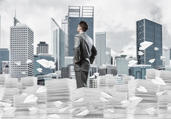 Takım elbiseli kendine güvenen işadamı arka planda şehir manzarası olan uçan kağıt uçaklar arasında evrak yığınının üzerinde duruyor. Karışık ortam.