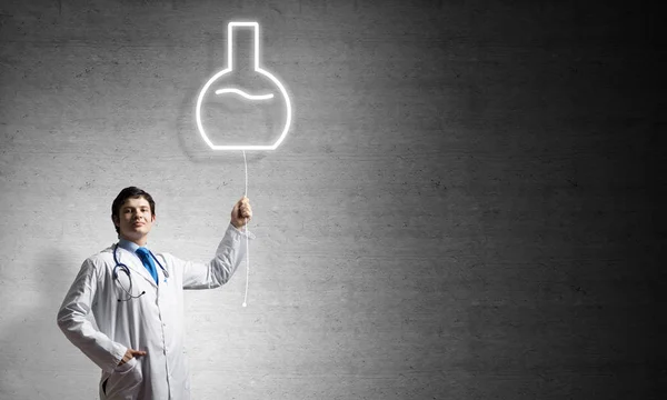 年轻自信的医生在白色医疗制服插带发光的小瓶符号在背景的深灰色墙壁站立在 — 图库照片