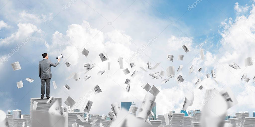 Businessman in suit standing among flying papers with speaker in hand and with skyscape on background. Mixed media.