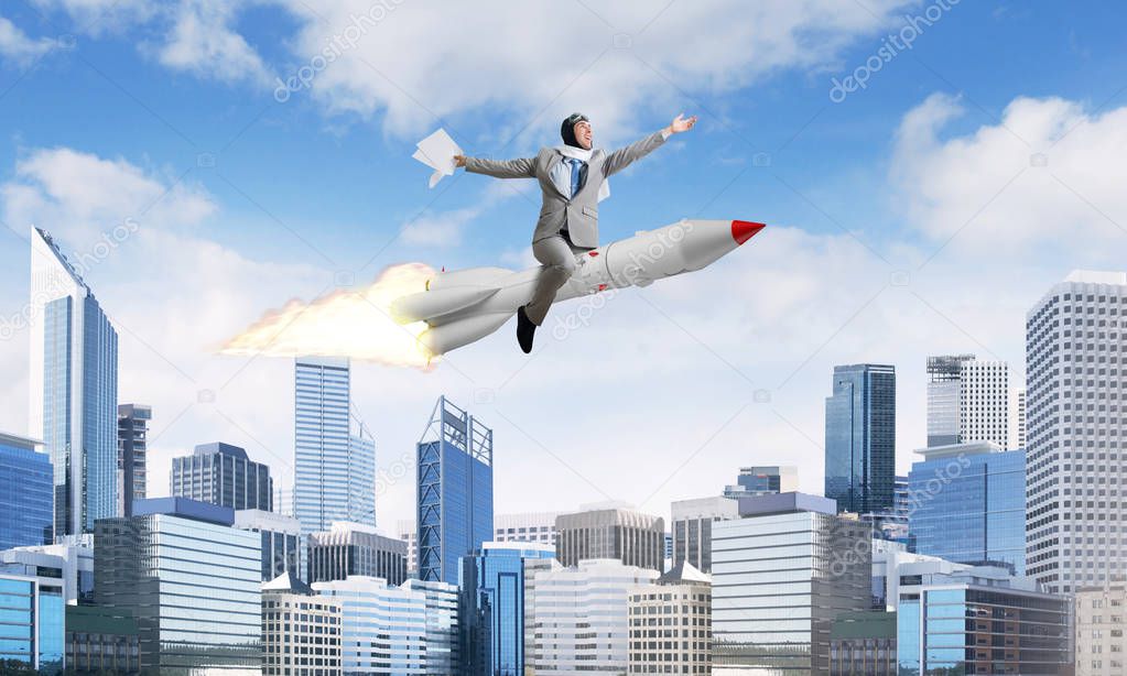 Conceptual image of young and happy businessman in suit flying on rocket with modern cityscape with skyscrapers and blue sky on background.