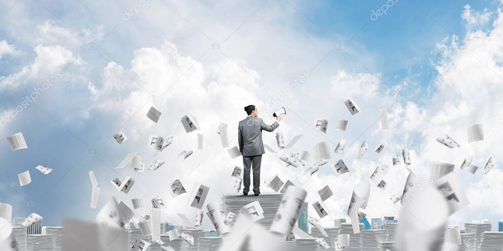 Businessman in suit standing among flying papers with speaker in hand and with skyscape on background. Mixed media.