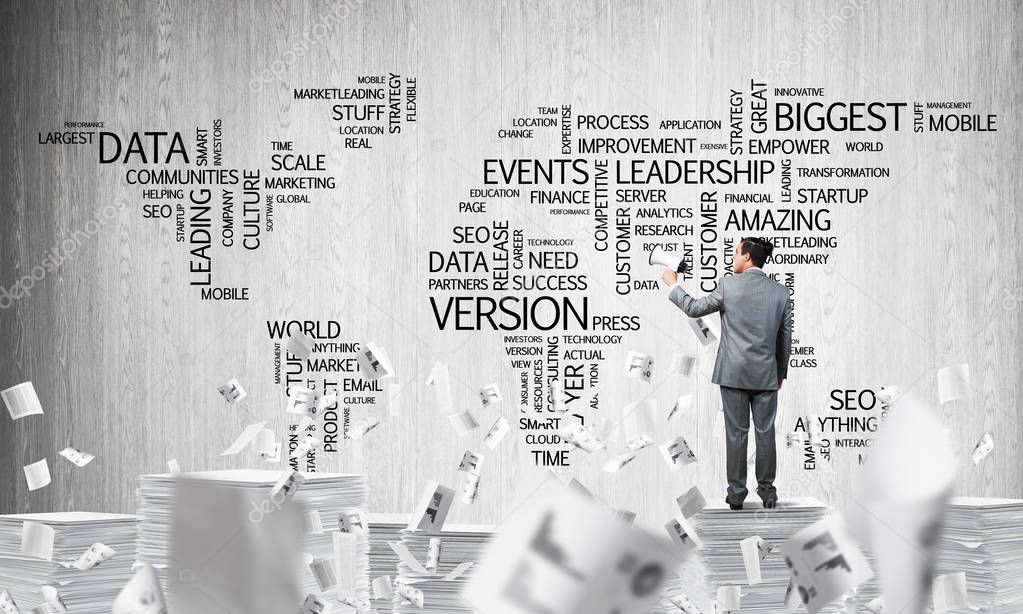 Businessman in suit standing among flying documents with speaker in hand with business-related terms in form of world map on background. Mixed media.