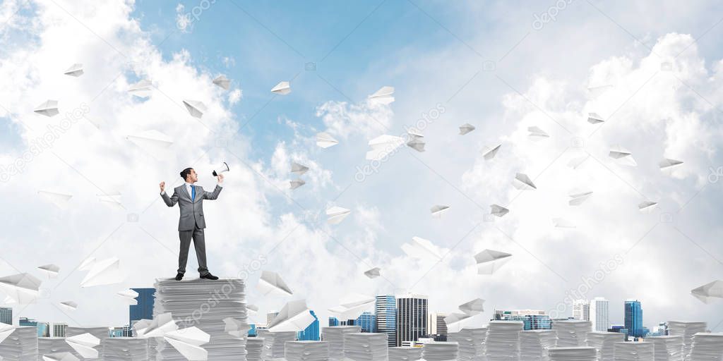 Businessman in suit standing among flying paper planes with speaker in hand and with skyscape on background. Mixed media.