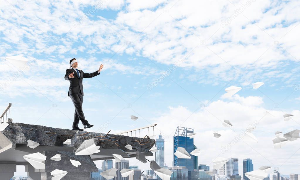 Businessman walking blindfolded among flying paper planes on concrete bridge with huge gap as symbol of hidden threats and risks. Cityscape view on background. 3D rendering.