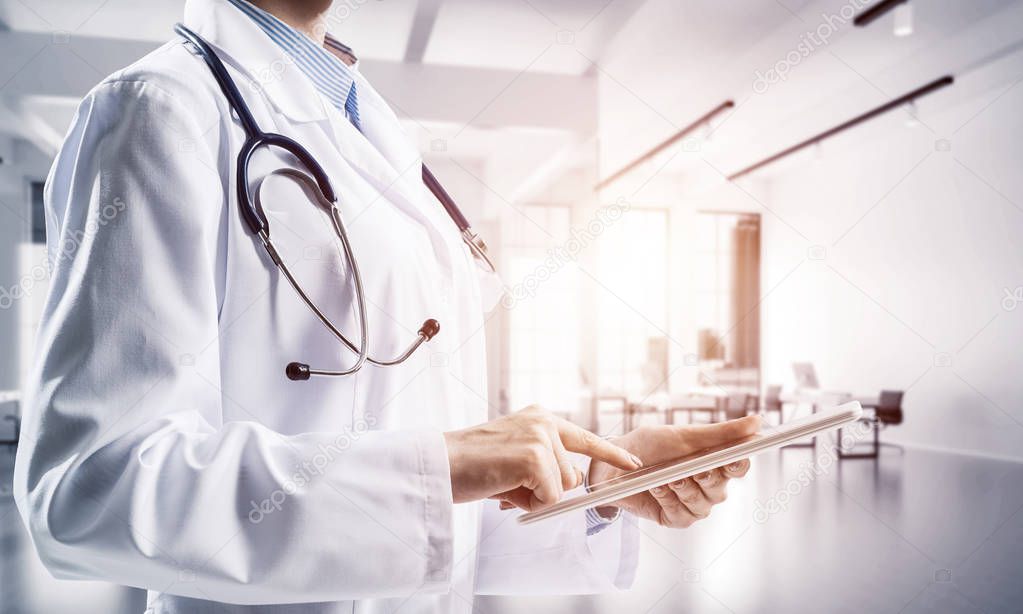 Close up of successful female doctor touching tablet with finger while standing indoors of white hospital building . Medical industry concept by means of confident doctor