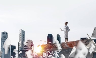 Kırık köprünün kenarında, arka planda şehir manzarası olan uçan kağıtların arasında duran beyaz üniformalı genç kadın doktor. Tıp endüstrisi konsepti