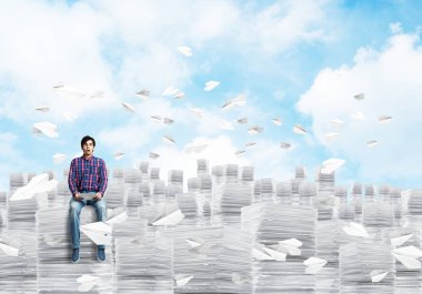 Casual giyim belgeler arasında kağıt uçaklar ile Clear gökyüzü arka plan üzerinde uçan yığını üzerinde oturan genç adam. Karışık teknik.