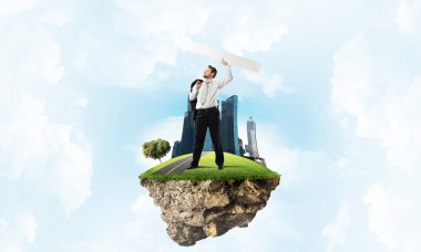 Kendine güvenen ve takım elbiseli genç işadamının uçan adada arka planda bulutlu bir gökyüzü varken havaya büyük beyaz bir ok attığı yatay bir çekim..