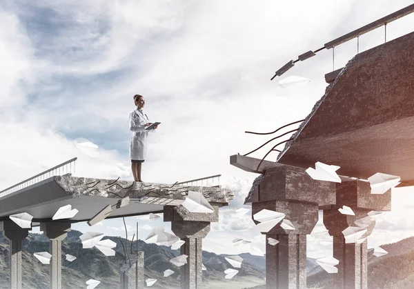 Professional medical industry employee using tablet while standing among flying paper planes at the edge of broken bridge with landscape view on background. Medical industry concept