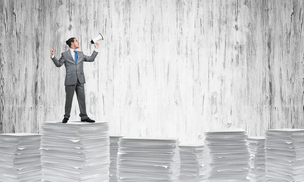 手の背景に灰色の壁とスピーカーと書類の山に立っているスーツのビジネスマン ミクスト メディア — ストック写真