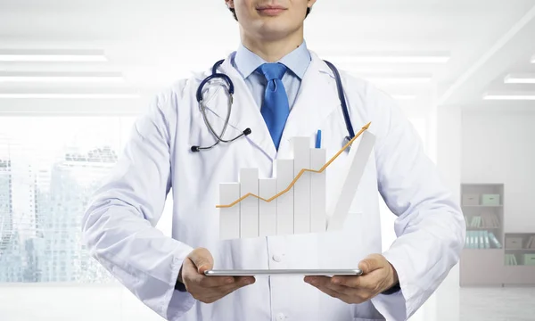 Cropped image of young doctor in sterile medical suit standing with tablet and graphical chart in his hands against bright office view on background.