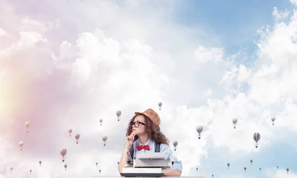 年轻和美丽的女作家在帽子和眼镜使用打字机 而坐在桌子上飞行轻飞行器和多云影像背景 — 图库照片
